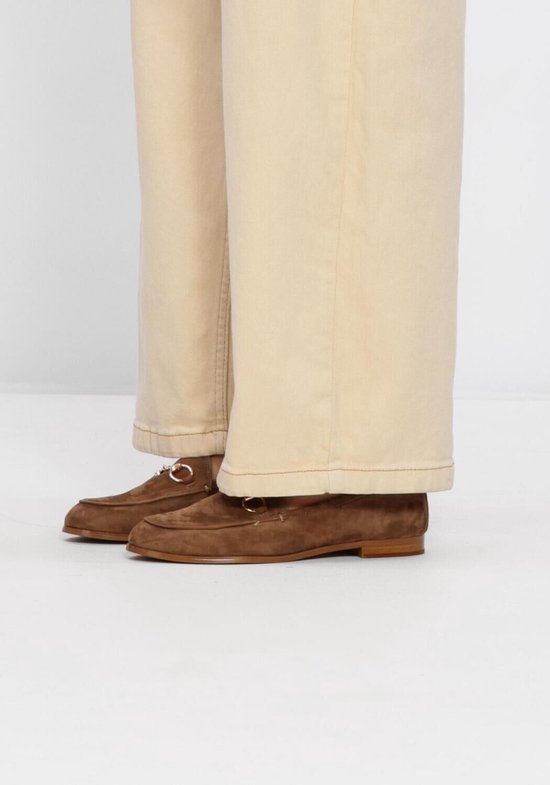 Een persoon draagt bruine suède loafers van Stefano Lauran met gouden bitdetail, met wijde beige broek.