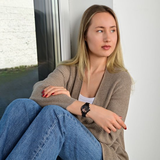 Jong blond meisje met jeans en vest zit bij een raam en draagt een zwart horloge met sterren.