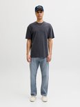 Man met donkere t-shirt, blauwe jeans en pet, staand met handen in de zakken.