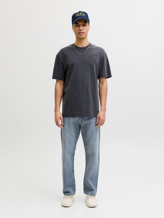 Man met donkere t-shirt, blauwe jeans en pet, staand met handen in de zakken.