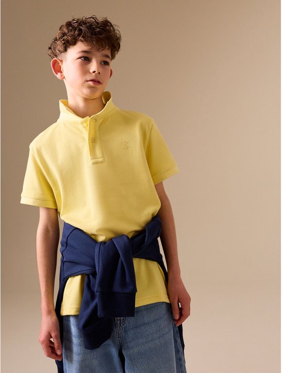 Jongen met krullend haar draagt een lichtgeel poloshirt met korte mouwen en een donkerblauwe trui om zijn middel.