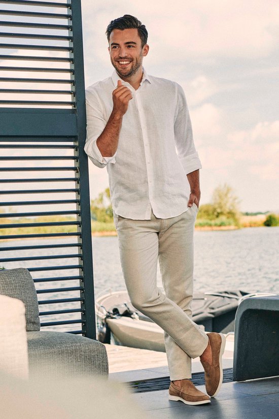 Man in een witte linnen blouse en beige broek, staand bij een waterkant.