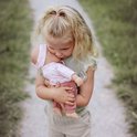 Een meisje met blond haar omhelst een pop met een roze outfit en hoofdband.