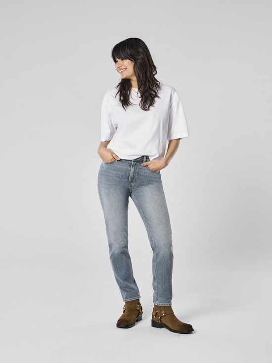 Vrouw met donker haar in een witte T-shirt en grijze spijkerbroek, lachend.