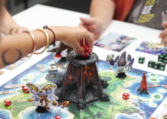 Close-up van het bordspel 'King of Monster Island' met een hand die een dobbelsteen op de vulkaan legt.