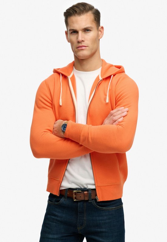 Homme aux bras croisés portant un sweat à capuche zippé Superdry orange, un t-shirt blanc et un jean.