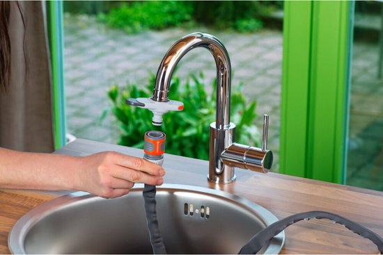 Een hand met een grijze tuinslang met oranje koppeling wordt aangesloten op een glanzende chromen kraan boven een rvs spoelbak.