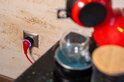 Blauwe Shelly Plug S met rode kabel in een stopcontact, naast een rode koffiemachine.