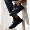 Zwarte lage werksneakers met veters, mesh en robuuste zolen, gedragen tegen een muur.
