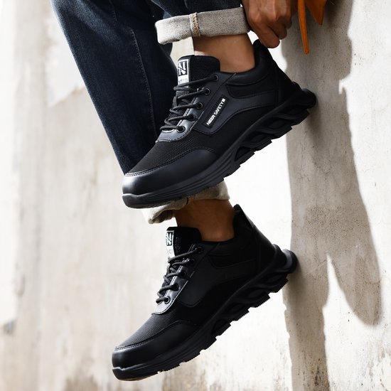 Zwarte lage werksneakers met veters, mesh en robuuste zolen, gedragen tegen een muur.