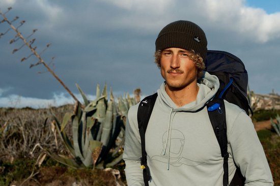 Man met krullend haar en Nomad-muts draagt een mintgroene hoodie en rugzak in de natuur.