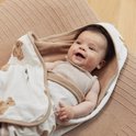 Bébé souriant, drapé dans une cape de bain blanc cassé avec imprimé ours en peluche et bordure beige. La cape a une doublure en tissu éponge marron.