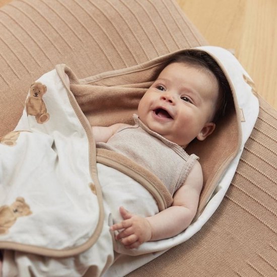 Bébé souriant, drapé dans une cape de bain blanc cassé avec imprimé ours en peluche et bordure beige. La cape a une doublure en tissu éponge marron.