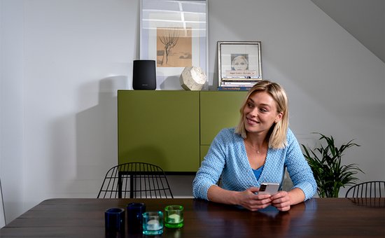 Zwarte Harman Kardon speaker op een groene kast, met een vrouw die op haar telefoon kijkt.