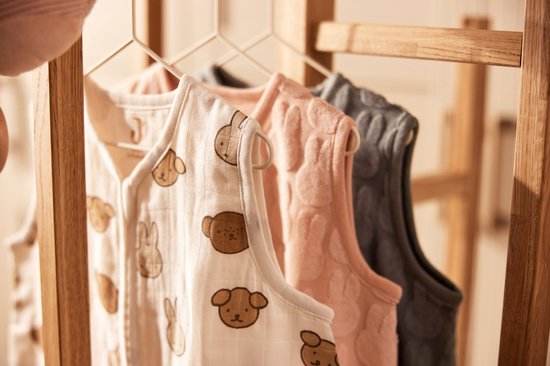 Drie mouwloze slaapzakken aan een houten rek. De voorste is wit met een Miffy print, de middelste is roze en de achterste is grijs.