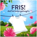 Un T-shirt blanc sèche sur une corde à linge dans un champ de fleurs jaunes et d'herbe verte. Des fleurs roses et des pétales flottent dans l'air.
