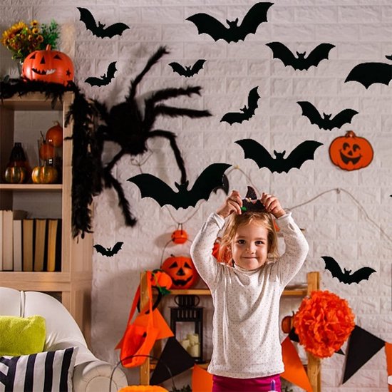 Een klein meisje met een witte trui en roze broek houdt een halloweenhoofddeksel omhoog, omringd door zwarte vleermuisdecoraties en pompoenen.