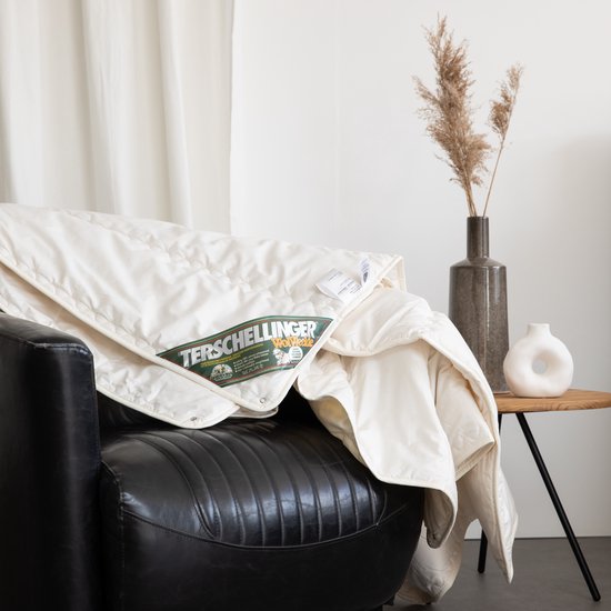 Wit, gewatteerd dekbed van Terschellinger Wolweelde op een zwarte fauteuil. Labels zichtbaar, met gedroogde grassen op de achtergrond.