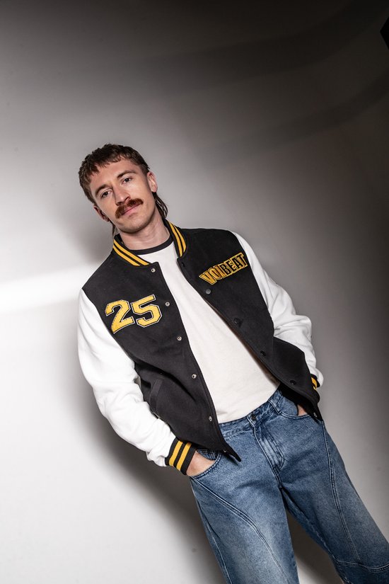 Model met baseballjack in zwart met witte mouwen, gele opdruk '25' en 'VOLBEAT'.
