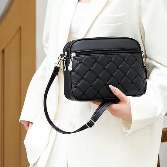 Zwarte gewatteerde crossbody tas met geborduurde hartjes, vastgehouden door een vrouw in een wit jasje.
