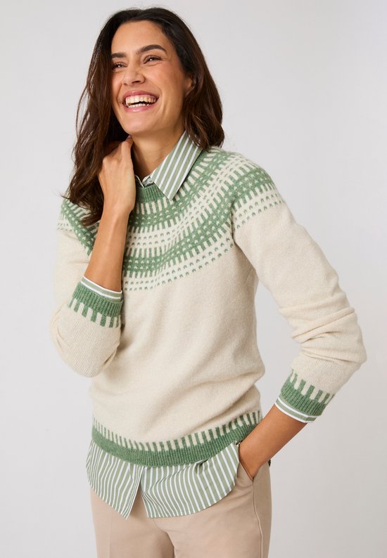 Lachende vrouw draagt een crèmekleurige jacquardtrui met groen patroon over een gestreept hemd en beige broek.
