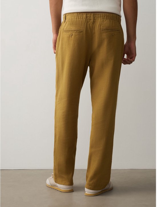 Achterkant van okergele broek van linnenmix met elastische tailleband, riemlussen en twee paspelzakken met knoop.