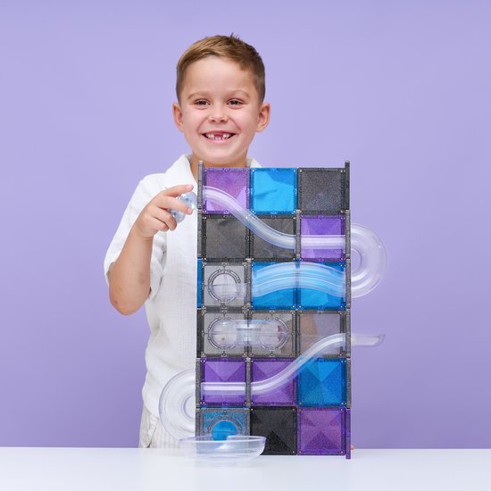 Een lachende jongen met een witte polo houdt een knikker vast, naast een hoge Connetix knikkerbaan van paarse, blauwe en grijze tegels.