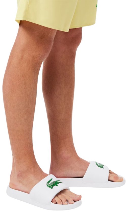 Witte Lacoste badslippers met groene krokodil op de band. Gedragen door een model met gele shorts.