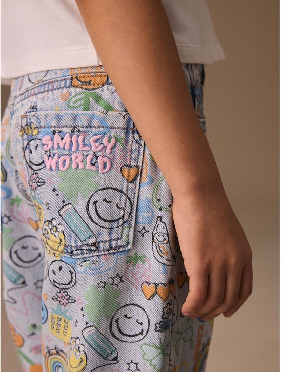 Close-up van een lichtblauwe spijkerbroek met all-over print van Smiley World-figuren en 'Smiley World'-logo op de achterzak.