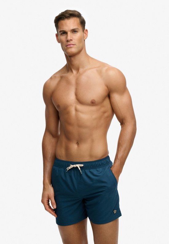 Un homme porte un short de bain bleu foncé avec logo, debout.