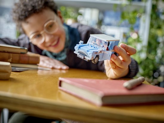 Une personne jeune tient une Ford Anglia Lego bleu clair avec Harry Potter et Ron Weasley en figurines dans la voiture. Des livres sont posés sur une table en bois.
