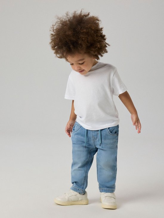 Jongen met krullend haar draagt een lichtblauwe spijkerbroek en witte sneakers.