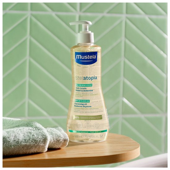 Gele doucheolie in een Mustela-fles met witte pomp, tegen een achtergrond van mintgroene tegels en handdoeken.