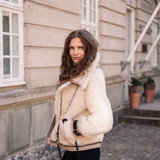 Femme aux longues boucles brunes, porte une veste en shearling crème avec des accents en cuir noir, des fermetures éclair et des boucles.