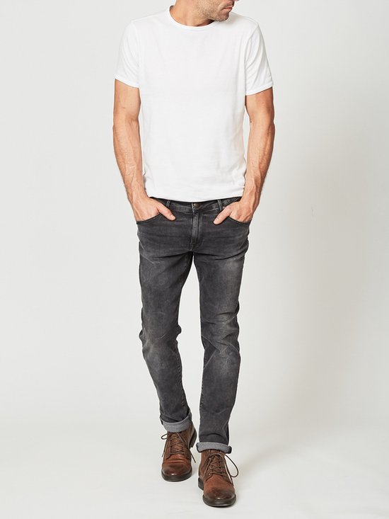 Man met de handen in zwarte slim fit jeans met opgerolde broekspijpen, gedragen met wit T-shirt en bruine schoenen.