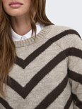 Close-up van beige gebreide trui met donkerbruin chevronpatroon, gedragen over een wit overhemd.