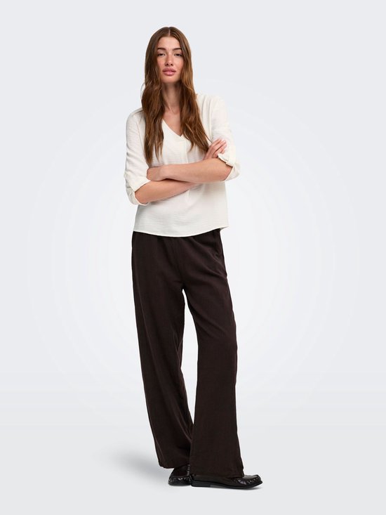 Model met gekruiste armen, draagt een witte blouse en bruine wijde broek.
