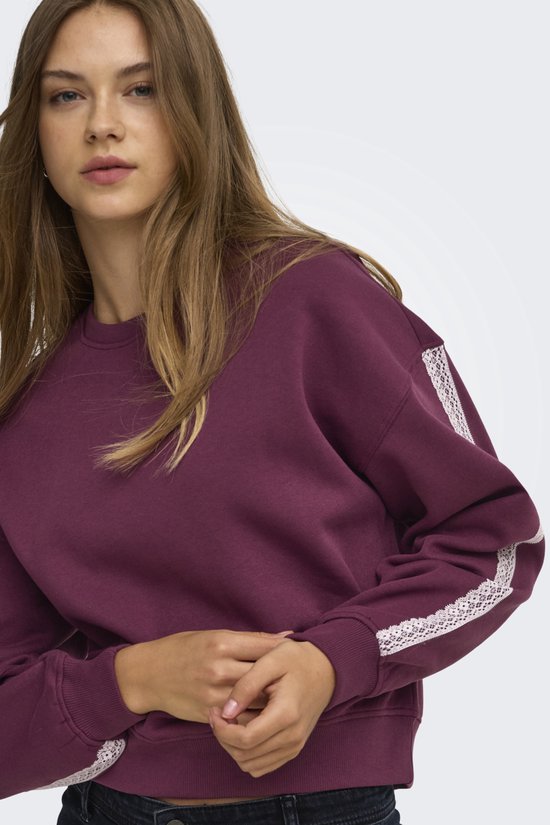 Model in paarse sweater met witte kanten mouwdetails, met de handen gevouwen in de taille.
