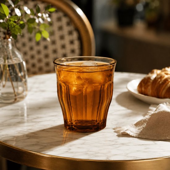 Une main tient un verre Duralex Picardie ambré avec des glaçons, du citron vert et une boisson.