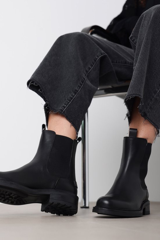 Zittend model draagt zwarte leren Chelsea boots bij een grijze jeans met rafels.
