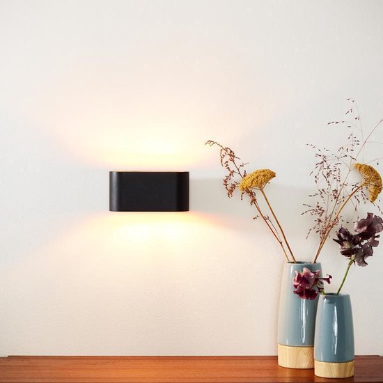 Zwarte XERA wandlamp, ovale vorm, met gouden binnenkant, boven een houten meubel met vazen.