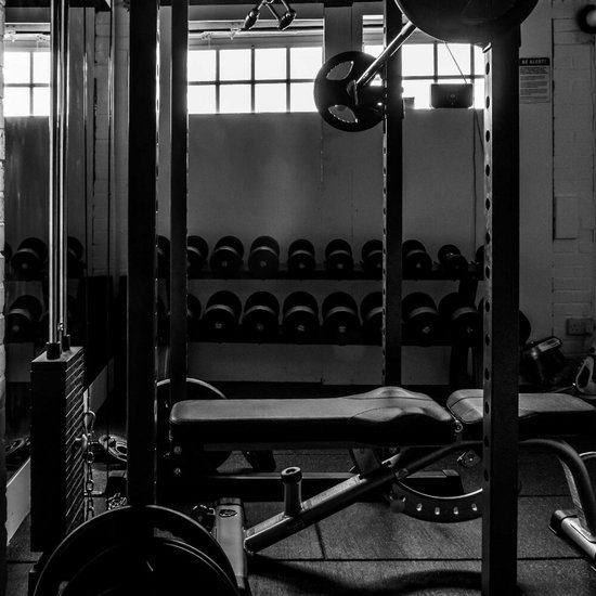 Zwart-wit foto van een fitnessruimte met een krachtstation, gewichtbank en rek met dumbbells voor een raam.