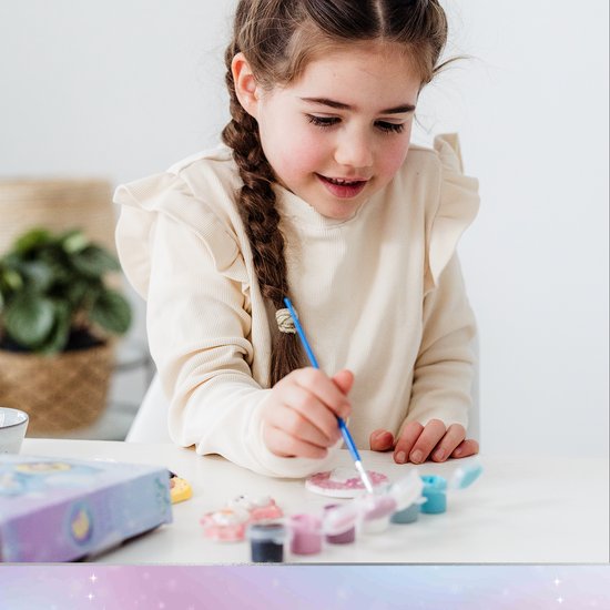Jong meisje met gevlochten haar schildert geconcentreerd Disney Frozen gipsfiguren aan een tafel.