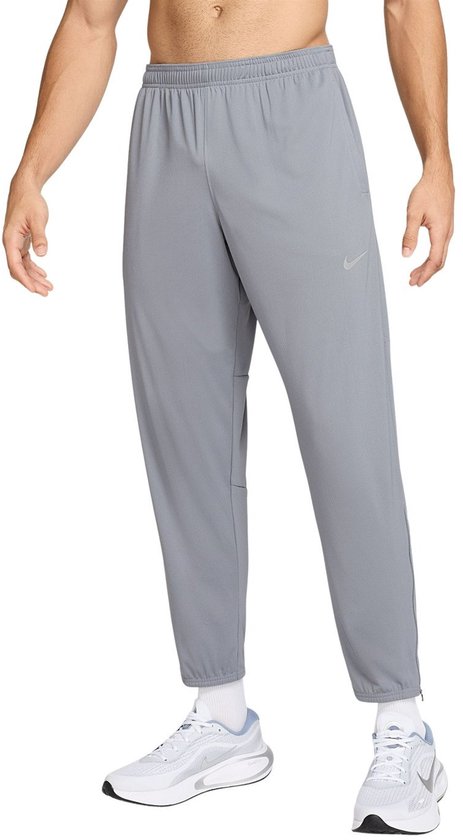 Man in een grijze Nike trainingsbroek met reflecterend logo, staand op witte sneakers.