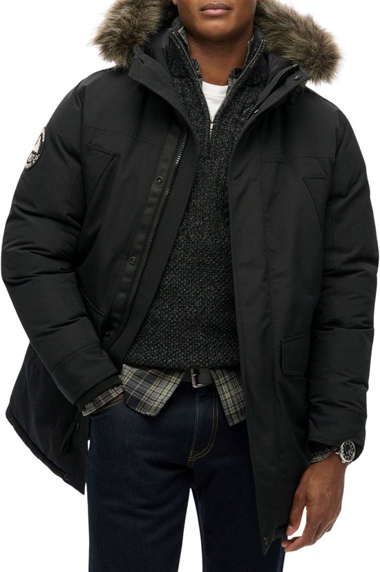 Man met bruine huid, gekleed in een zwarte Superdry parka met bruine faux fur capuchon, een grijze trui en geruite blouse.