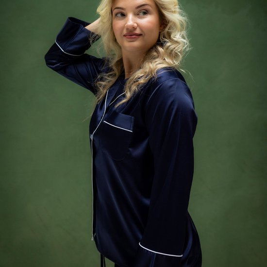 Model draagt een donkerblauwe satijnen pyjamajas met witte biesjes, die haar hand door haar haar haalt.