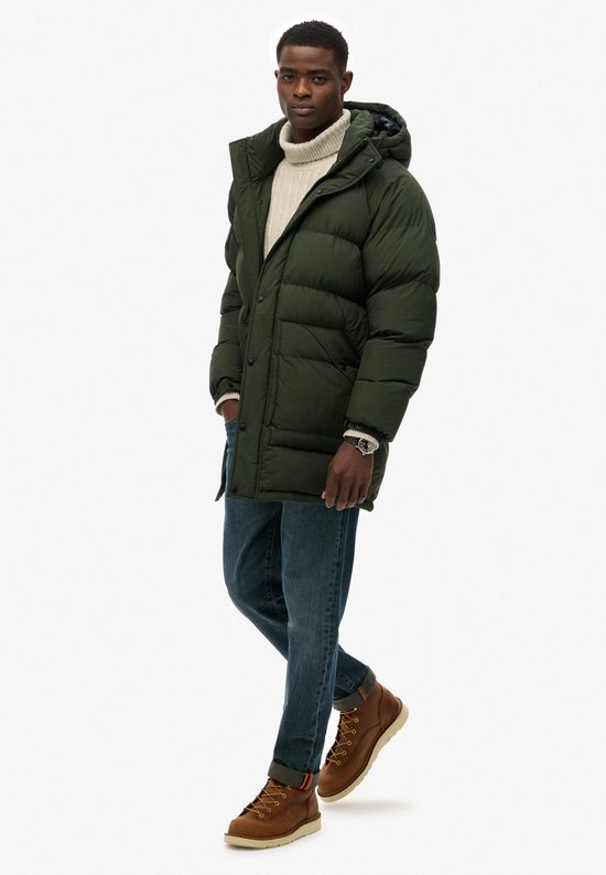 Groene lange gewatteerde parka met capuchon, gedragen door een man, staand.