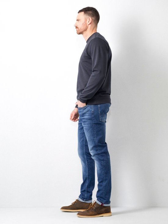 Man in een donkergrijs shirt en blauwe jeans, staand in profiel. Hij draagt bruine schoenen.