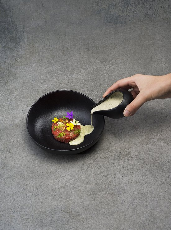 Een hand giet saus uit een zwarte sauskom over een tartaar in een zwarte schaal.