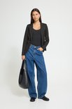 Model met lange zwarte blazer, één knoop sluiting, gedragen met een zwarte top, jeans en zwarte tas.
